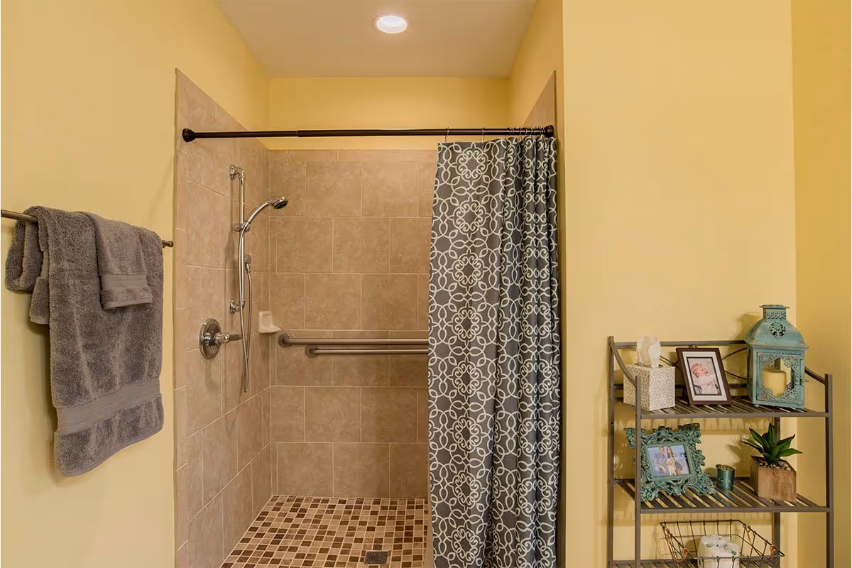 Walk-in tiled shower with a patterned curtain, grab bar and towel rack in a yellow-painted bathroom with a decorative shelving unit.