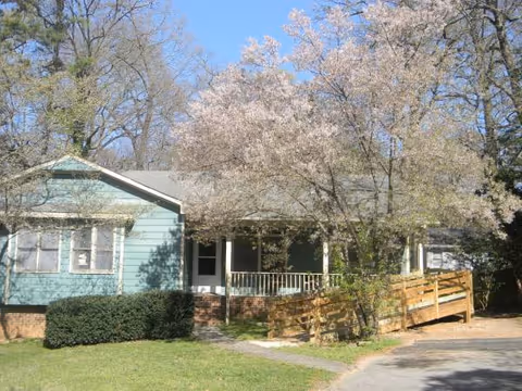 Single-story pastel blue senior living building with front porch, ramp, and blossoming trees.