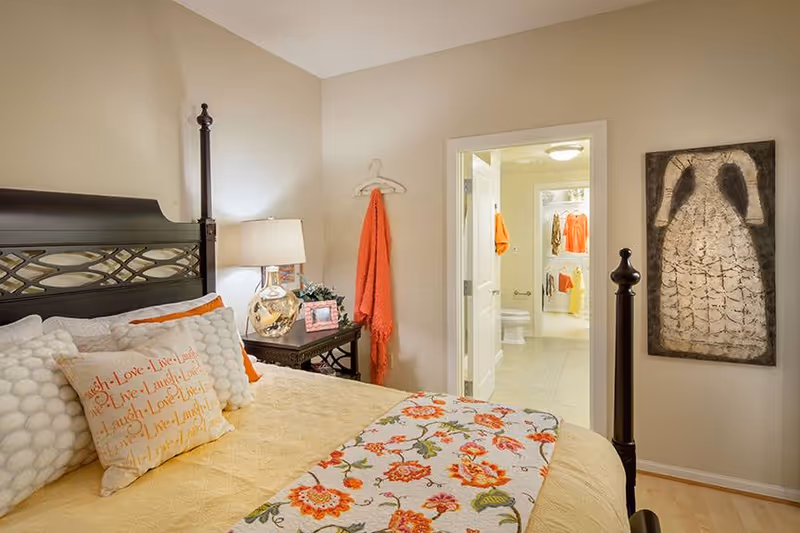 Cozy bedroom with a four-poster bed, floral bedding, nightstand with lamp, and an open door showing a bathroom/closet.