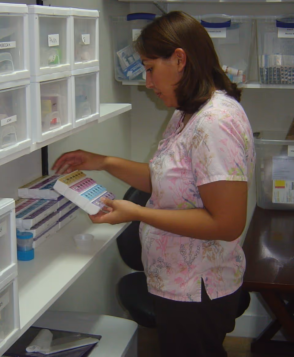 A woman in a floral scrub top is organizing or selecting medication from a shelf filled with labeled plastic containers and pill organizers in a medical or care facility setting.