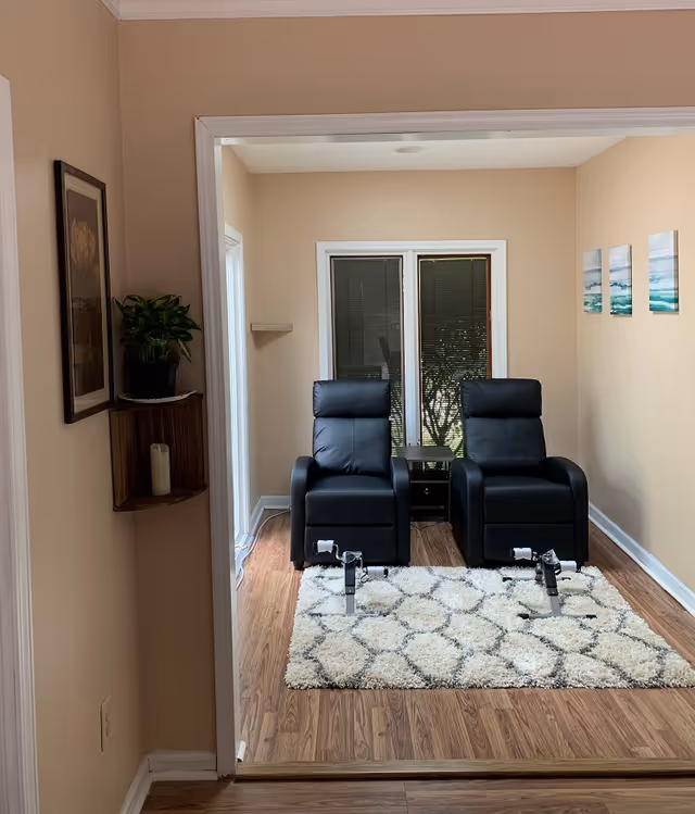Small sitting room with two black recliners on a patterned rug facing a window, with wall art and a potted plant nearby.