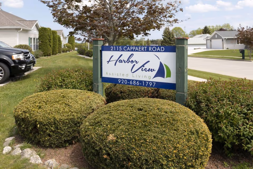 Outdoor view of a sign for Harbor View Assisted Living located at 2115 Cappaert Road, surrounded by trimmed bushes and greenery with residential houses and a parked black vehicle visible in the background.