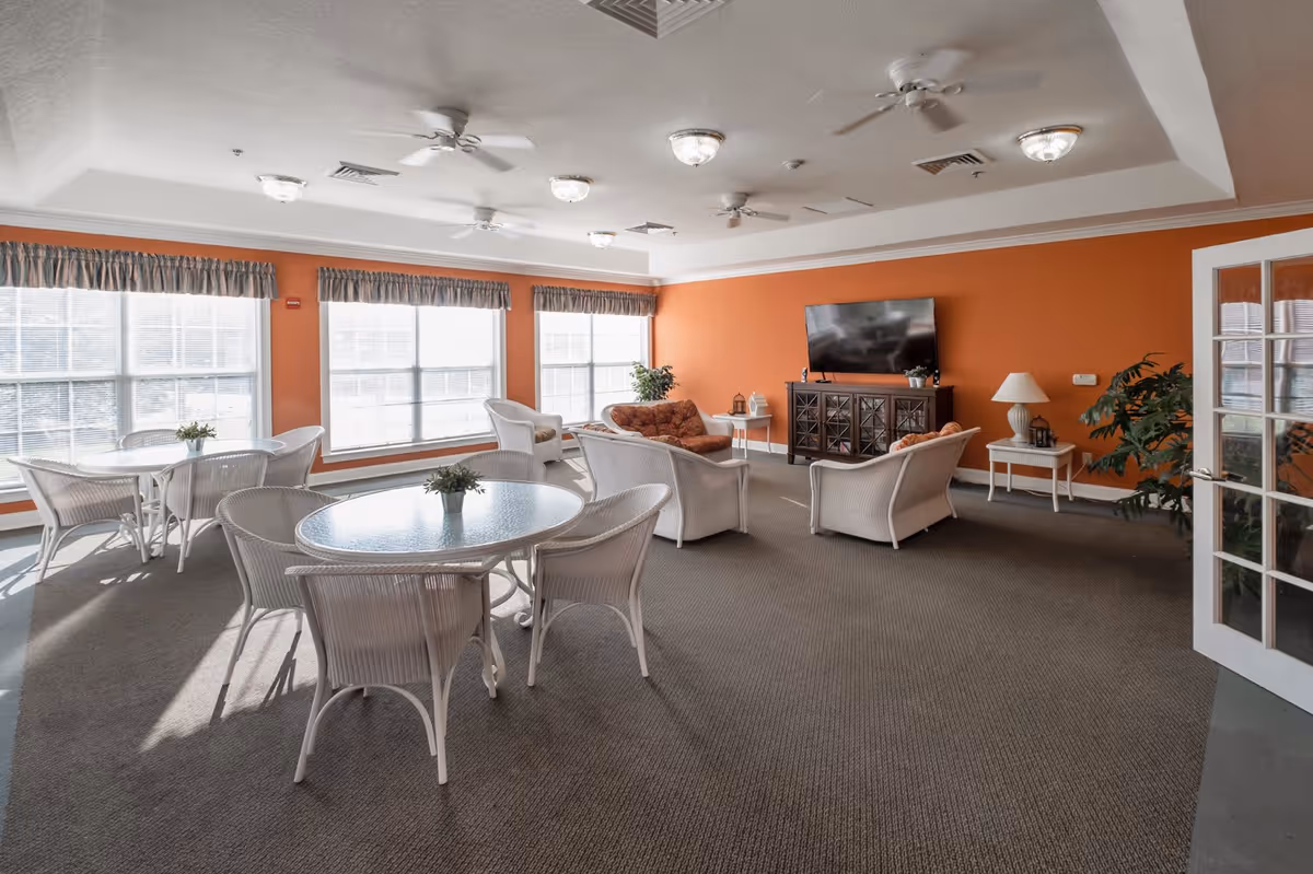 A bright and spacious common area with large windows letting in natural light. The room features white wicker chairs and round glass-top tables, a seating area with cushioned chairs and a sofa facing a flat-screen TV mounted on an orange accent wall. There are ceiling fans and light fixtures on the ceiling, and potted plants and lamps add decorative touches.