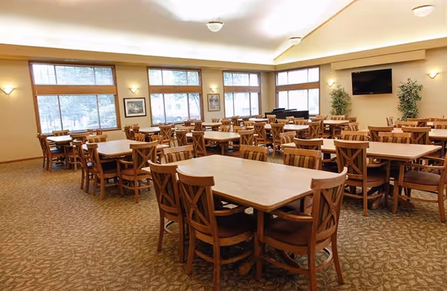 A spacious dining room with multiple wooden tables and chairs arranged neatly. Large windows allow natural light to fill the room. The walls are light-colored with sconces for additional lighting. A flat-screen TV is mounted on one wall, and there are two potted plants near it. The carpet has a patterned design.