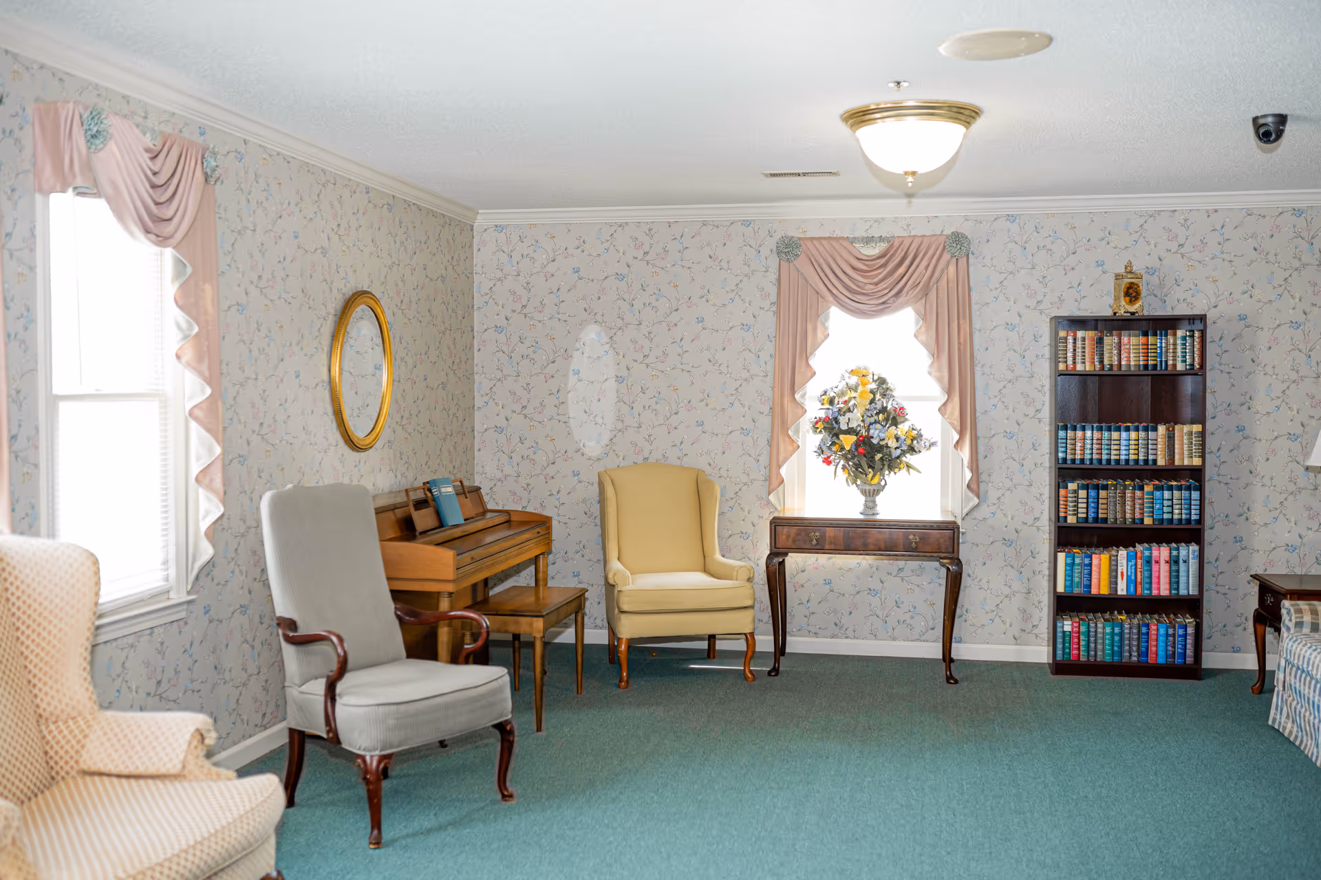 A cozy living room with floral wallpaper, green carpet, and a ceiling light. The room features a beige armchair, a gray armchair with wooden arms, a wooden piano with a bench, a small wooden table with a vase of colorful flowers in front of a window with pink drapes, and a tall bookshelf filled with books.