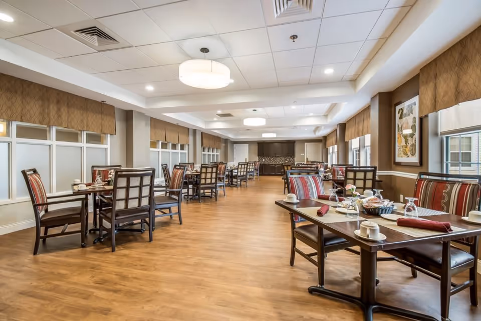 A spacious dining room with multiple tables and chairs arranged neatly. The tables are set with cups, plates, napkins, and glasses. The room features large windows with brown valances, wooden flooring, and ceiling lights providing bright illumination. Artwork is displayed on the walls.
