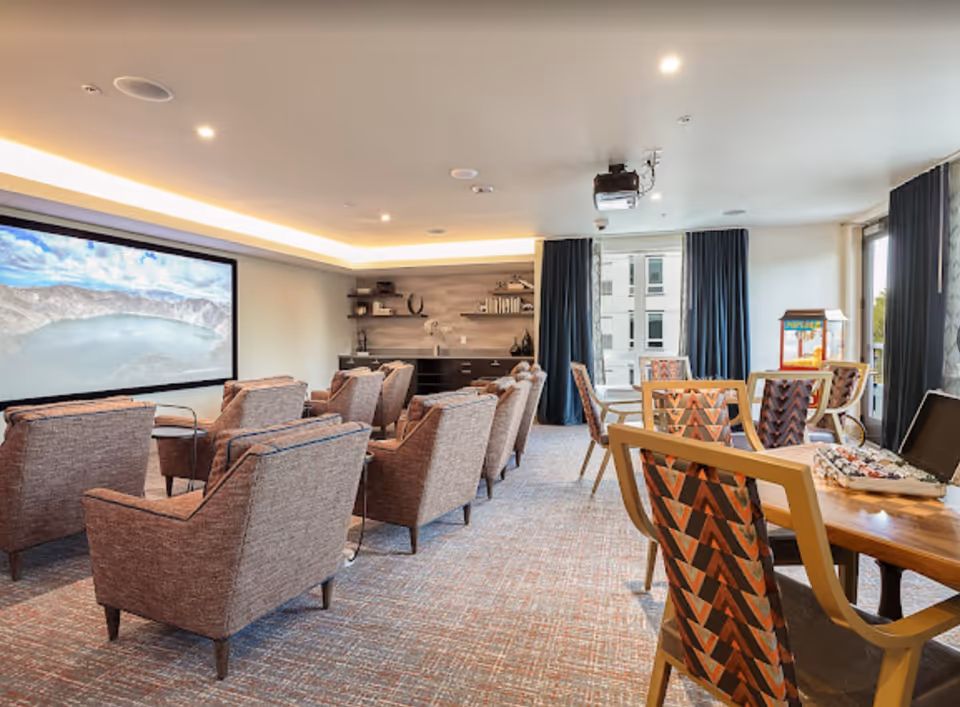 A spacious, well-lit common room with multiple comfortable armchairs arranged in rows facing a large screen on the wall. The room also features a table with patterned chairs, a popcorn machine, and large windows with dark curtains allowing natural light to enter.