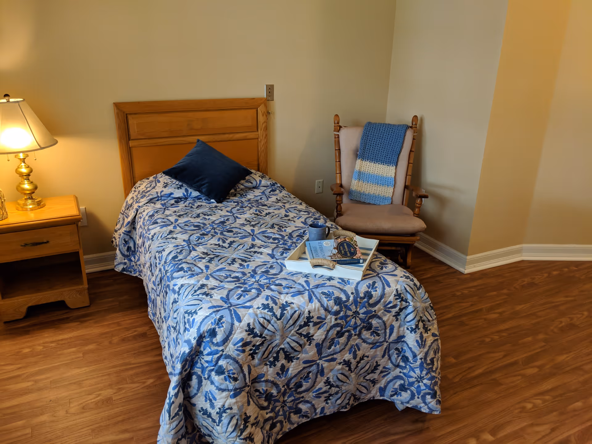 A single bed with a blue and white patterned quilt and a dark blue pillow. A wooden nightstand with a brass lamp is next to the bed. A wooden rocking chair with a beige cushion and a blue and white knitted blanket draped over the back is positioned in the corner. The floor is wood and the walls are painted a light beige color.