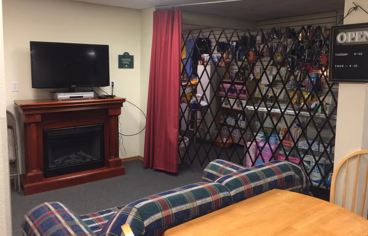 Interior view of a common area in a senior living facility with a plaid couch, a wooden table and chairs, a TV mounted above a wooden electric fireplace, and a gated general store stocked with various items behind a black metal lattice gate.