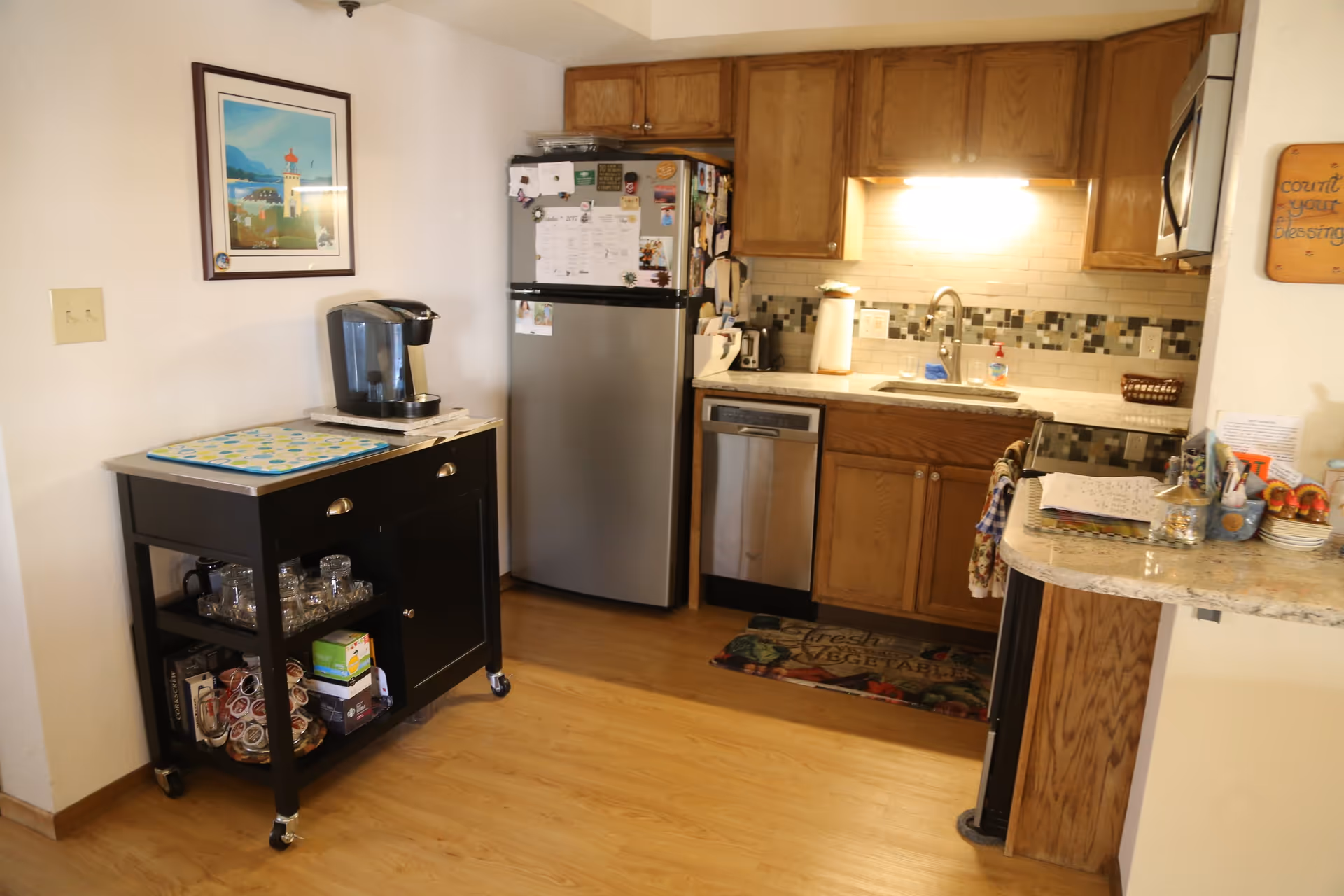 A small kitchen area with wooden cabinets, a stainless steel refrigerator, dishwasher, and microwave. There is a black rolling cart with a coffee maker and various items on it. The countertop has a sink with a faucet, paper towel holder, and soap dispenser. A framed picture hangs on the wall above the cart, and a decorative sign is mounted on the wall near the countertop.