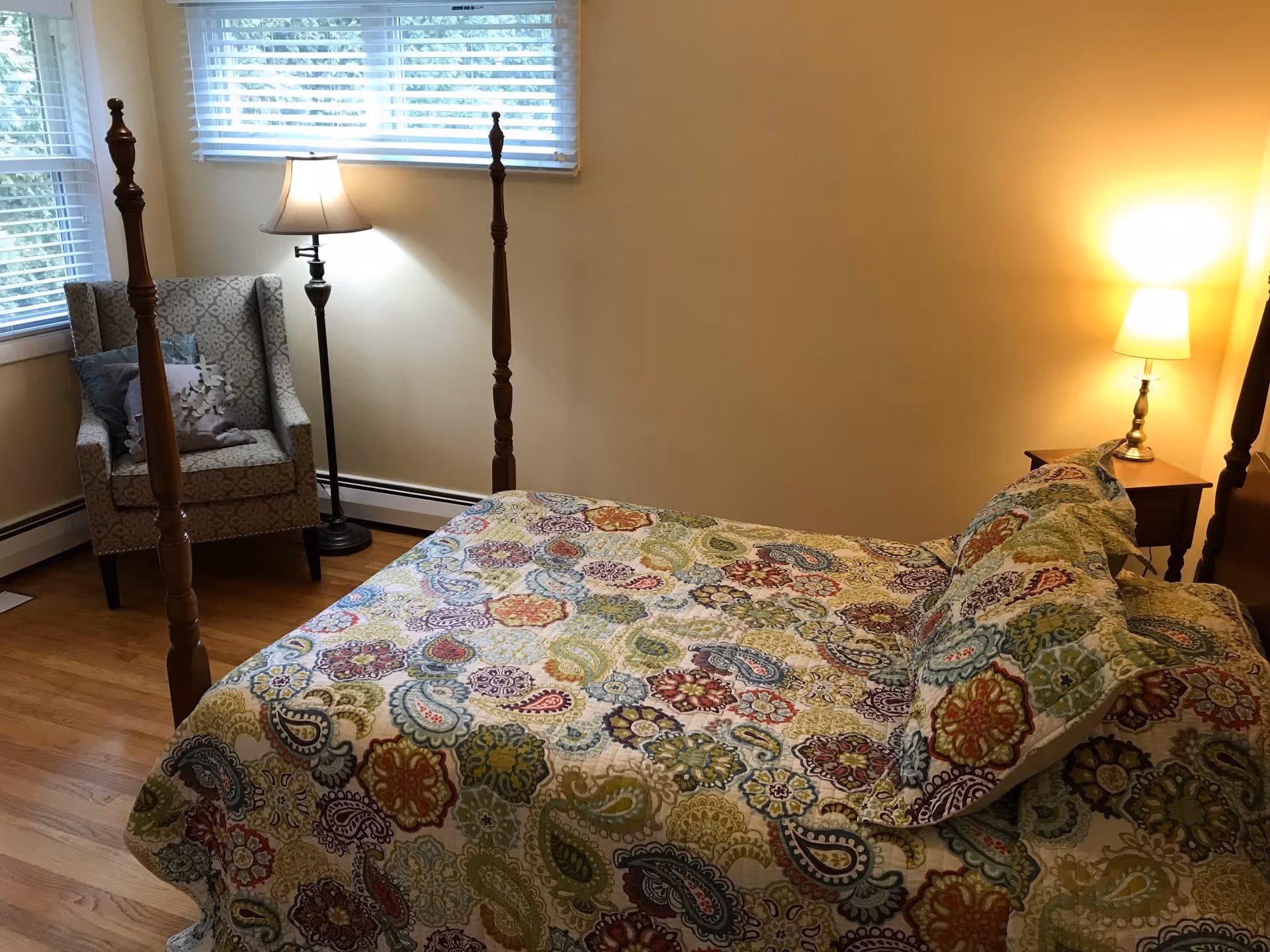 A cozy bedroom with a wooden four-poster bed covered in a colorful floral and paisley patterned quilt. There is a cushioned armchair with decorative pillows near two windows with white blinds. Two lamps, one floor lamp and one table lamp on a wooden nightstand, provide warm lighting. The room has light-colored walls and hardwood flooring.