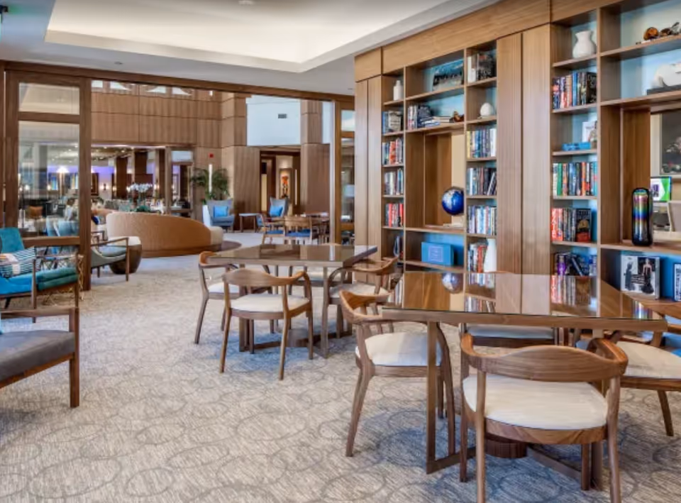A spacious and well-lit common area with wooden bookshelves filled with books and decorative items, several wooden tables with cushioned chairs, and additional seating including armchairs and sofas in the background. The room features a carpeted floor and a modern ceiling design with recessed lighting.