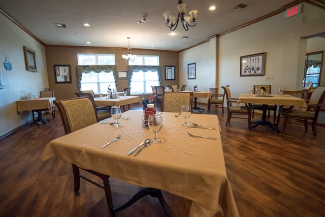 A dining room in Cedar Hill Senior Living with several tables covered in beige tablecloths, each set with glasses, silverware, and condiments. The room has wooden flooring, framed artwork on the walls, and windows with valances letting in natural light.