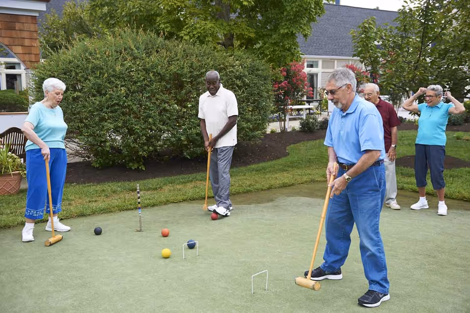 Five elderly people playing croquet on a green lawn outside a residential building, with bushes and trees in the background. Two men and one woman are holding croquet mallets, while two other people watch and cheer.