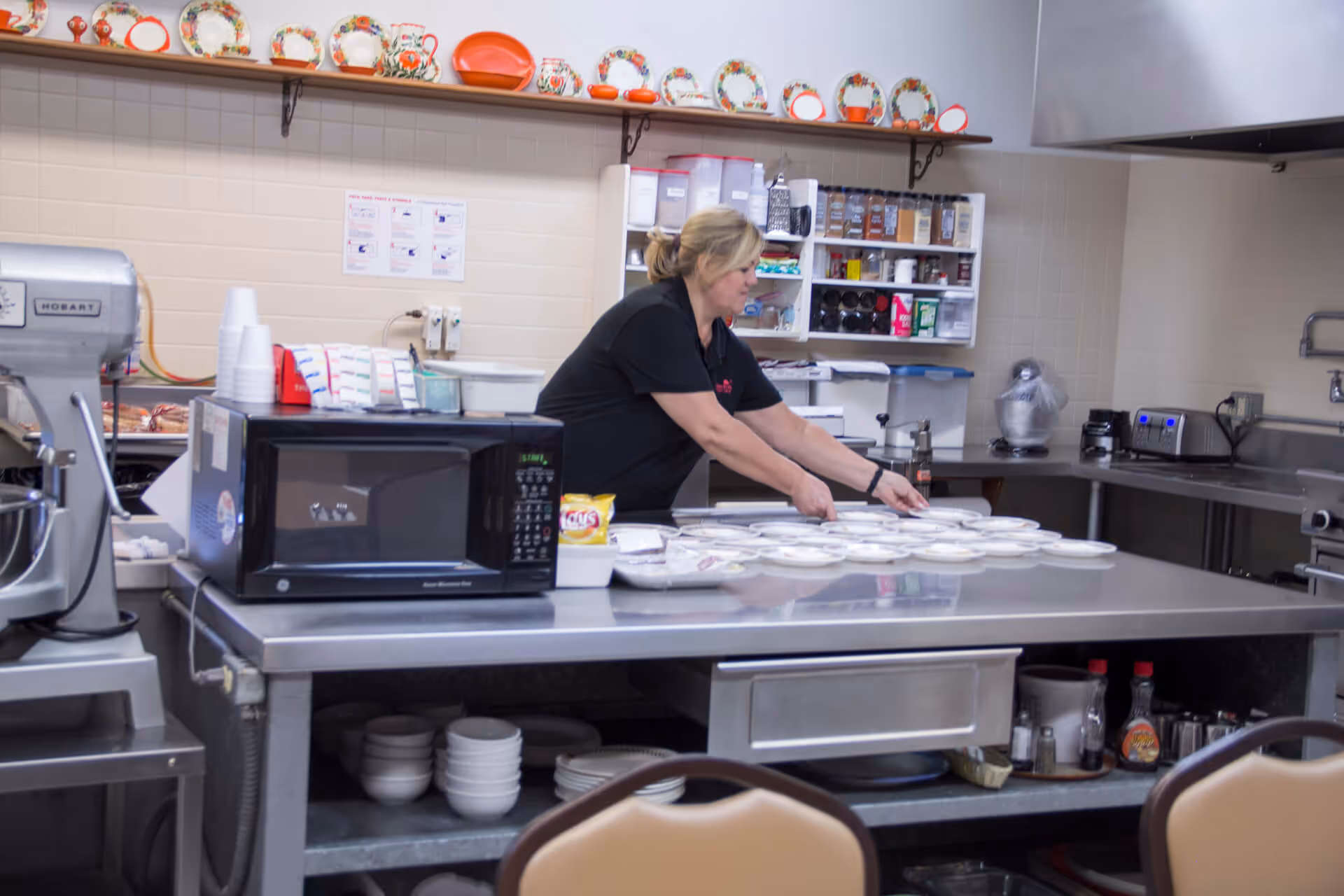 A woman in a black shirt is arranging plates on a stainless steel counter in a kitchen. The kitchen has various appliances including a microwave, a mixer, and shelves stocked with spices and containers. Decorative plates and teapots are displayed on a shelf above the counter.