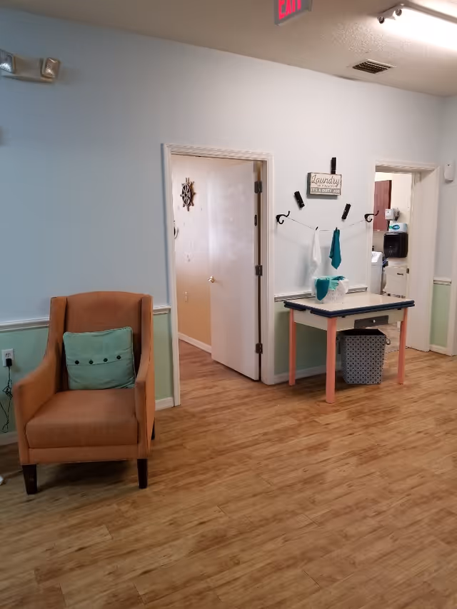 Interior view of a room with light blue and green walls, wooden floor, an orange armchair with a green pillow on the left, and a small table with a laundry basket and hanging towels on the right. Two open doorways lead to other rooms, one with a visible sink and paper towel dispenser. A sign above the table reads 'Laundry It's a Dirty Job'.