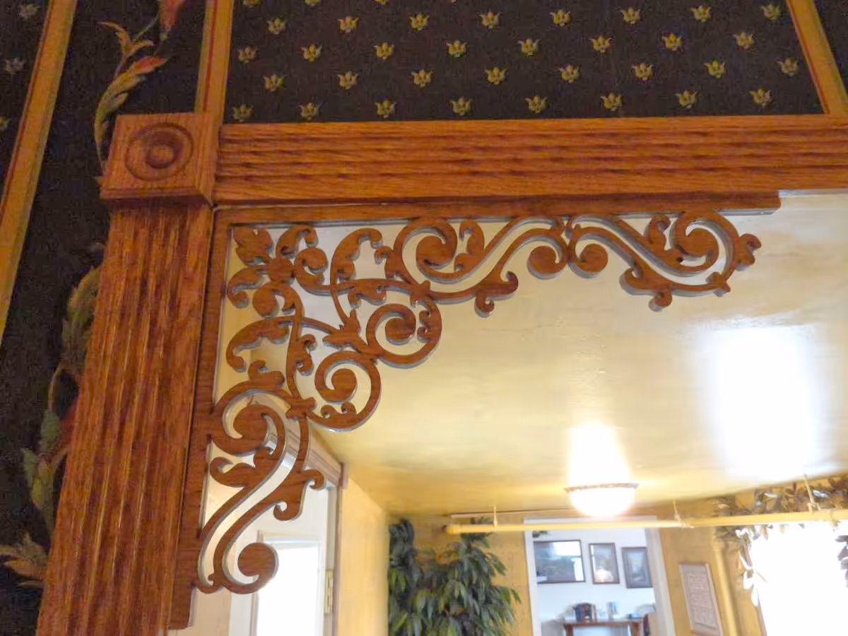Close-up view of an ornately carved wooden frame with intricate scrollwork and floral patterns, surrounding a mirror. The background shows part of a room with framed pictures on the wall, a plant, and a ceiling light fixture.