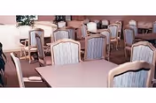 A dining room with multiple tables and chairs arranged neatly. The chairs have cushioned backs and seats with a striped fabric pattern. The room has soft lighting and a calm atmosphere.