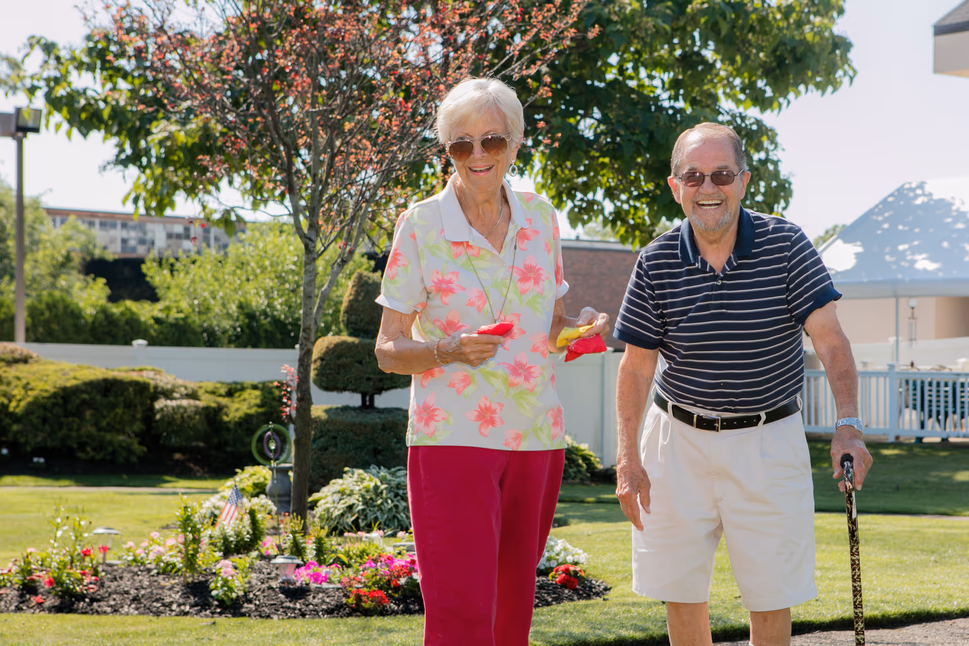 An elderly woman and man standing outside in a garden area. The woman is wearing sunglasses, a white shirt with pink floral patterns, and red pants, holding bean bags. The man is wearing sunglasses, a navy blue striped polo shirt, white shorts, and holding a cane. They are smiling and appear to be enjoying a sunny day at an assisted living facility.
