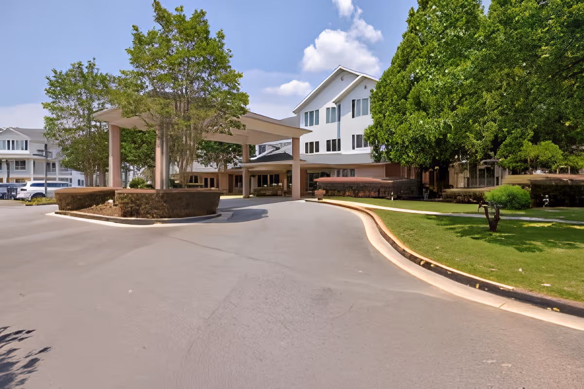 Driveway and covered entrance of a multi-story senior living building with trees and landscaped lawn.