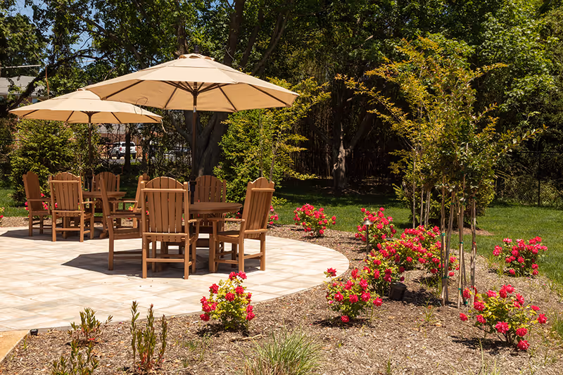 Outdoor patio area with wooden tables and chairs under beige umbrellas surrounded by green grass, trees, and blooming red flowers in a garden setting.