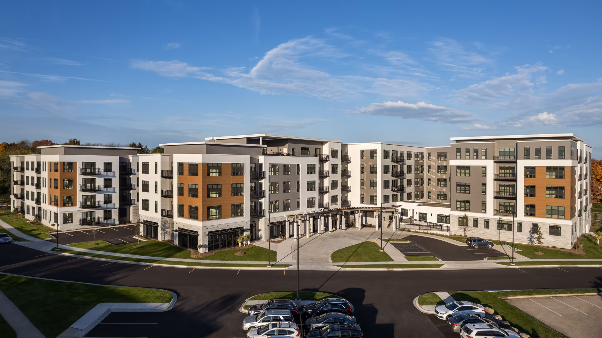 Exterior view of a modern multi-story senior living facility named Vista West with a large parking area in front, surrounded by green lawns and clear blue sky.