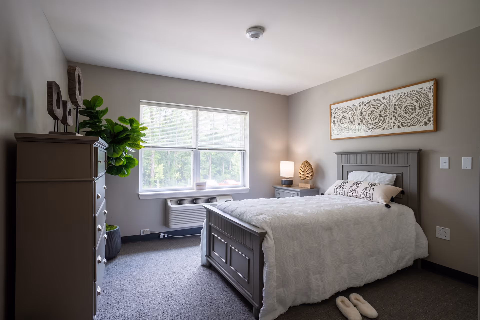 A neatly arranged bedroom with a gray wooden bed frame and white bedding. There is a decorative pillow on the bed, a nightstand with a lamp and a decorative leaf sculpture, a dresser with three wooden sculptures on top, and a large green potted plant next to the dresser. A window with blinds lets in natural light, and a framed artwork hangs above the bed. A pair of white slippers is placed on the carpeted floor near the foot of the bed.