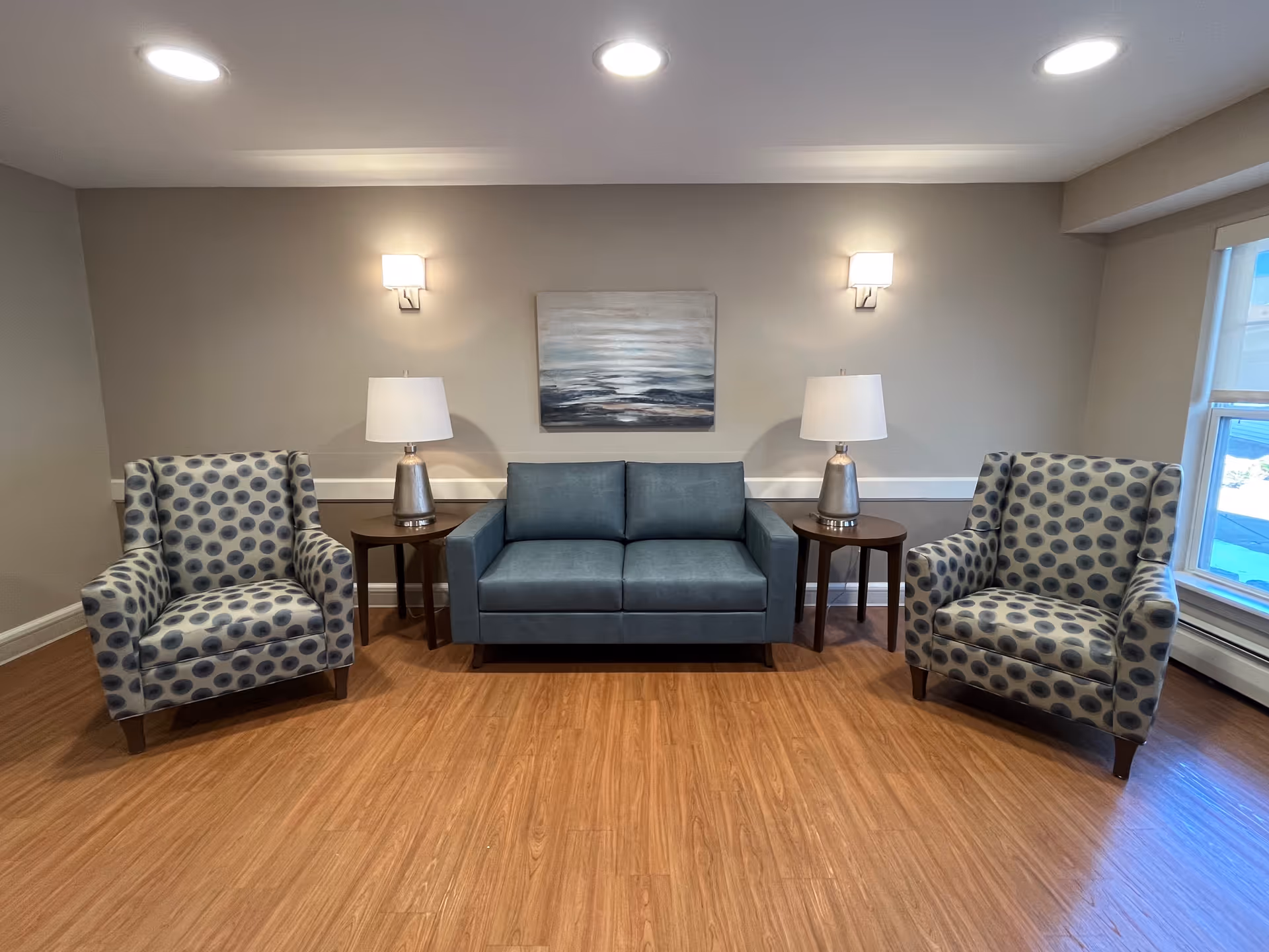 A small seating area with a blue loveseat centered between two patterned armchairs, side tables with lamps, and a painting on a beige wall.