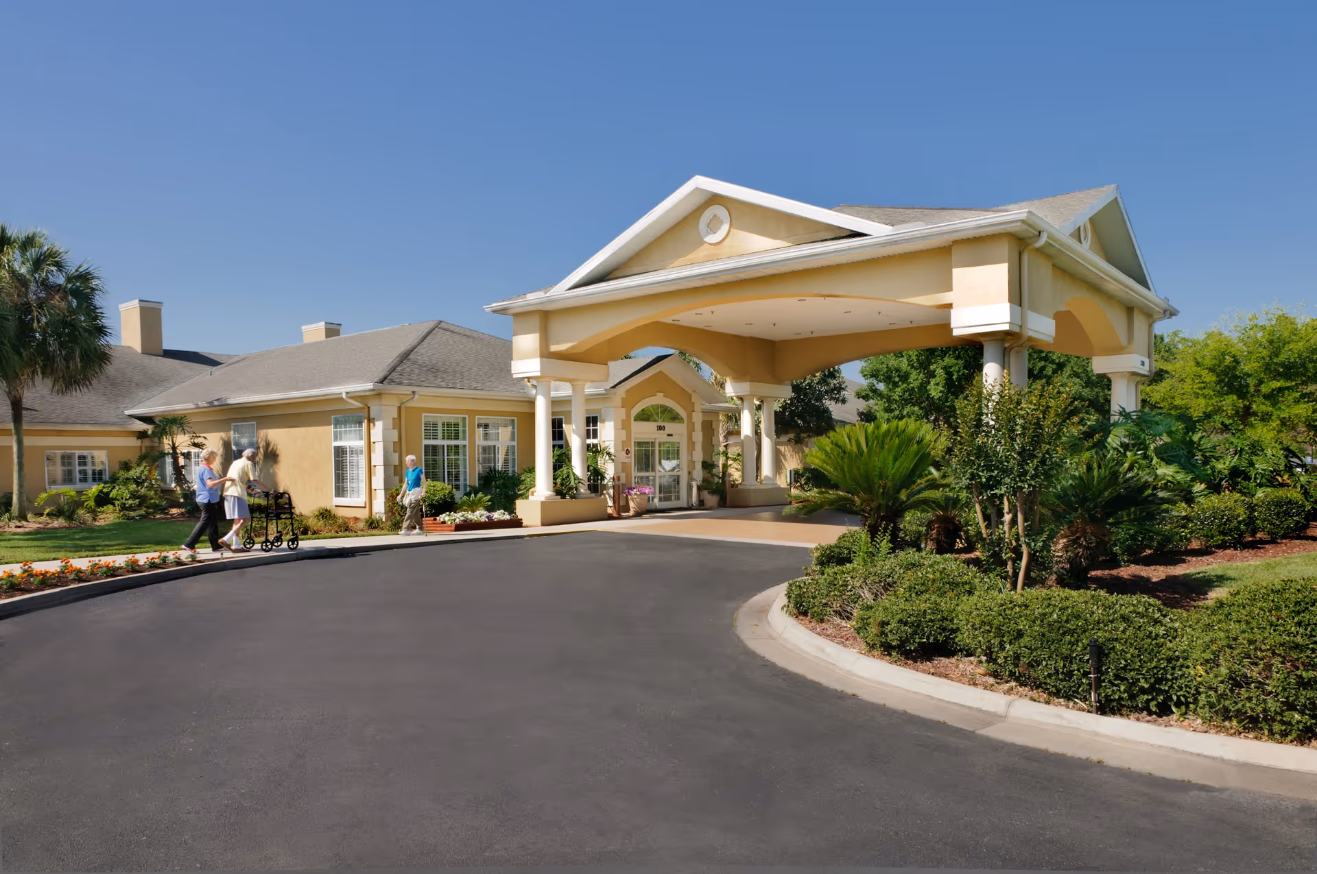 Front entrance of a yellow senior living facility with a covered porte-cochère, landscaped driveway, and people walking nearby.