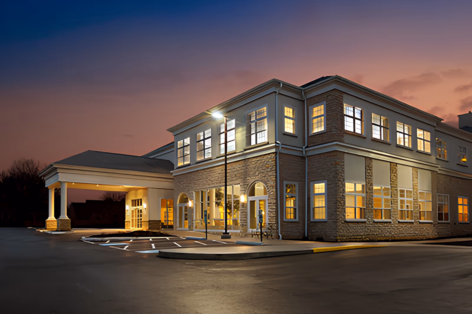 Front exterior of a two-story senior living building at dusk with illuminated windows and a covered entrance/porte-cochere.
