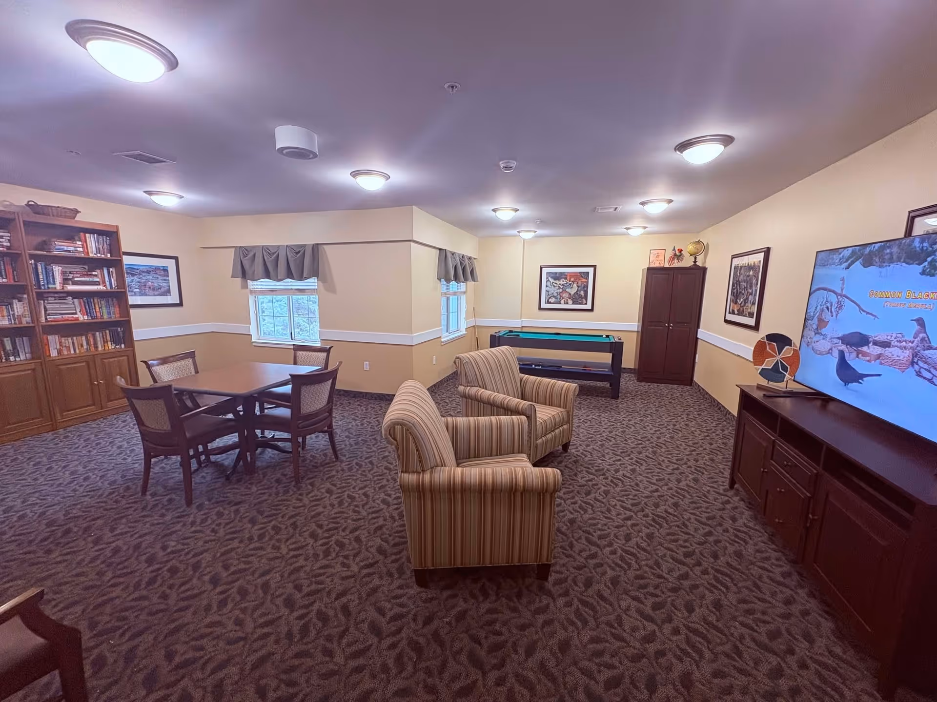 A cozy common room with patterned carpet and beige walls featuring a seating area with two striped armchairs facing a large flat-screen TV on a wooden cabinet. To the left, there is a wooden table with four chairs near two windows with valances. A bookshelf filled with books and framed artwork on the walls are also visible. In the back corner, there is a foosball table and a tall cabinet.