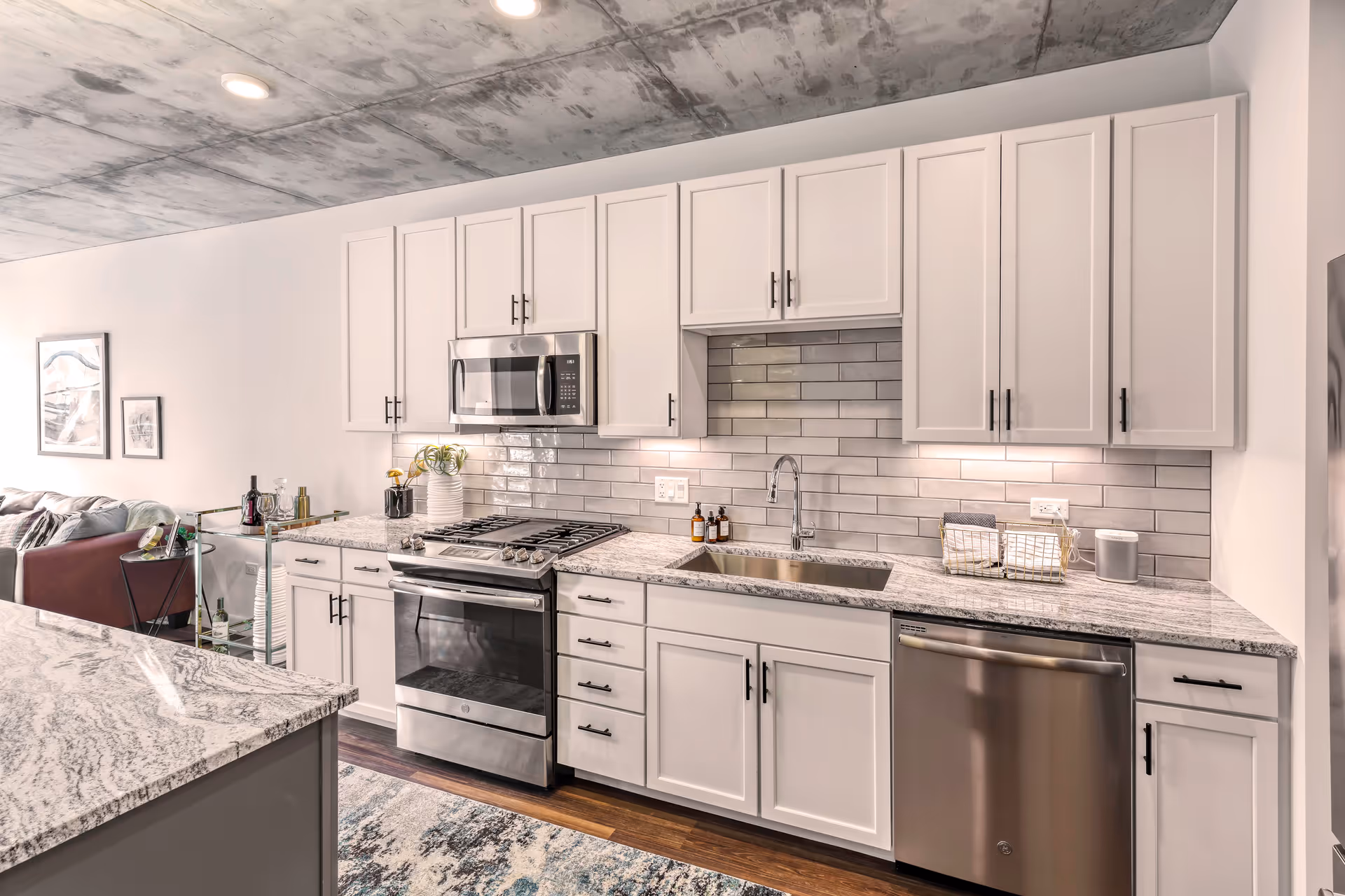 Modern kitchen with white cabinets, stainless steel appliances including a microwave, oven, and dishwasher, a granite countertop, and a subway tile backsplash. The kitchen opens to a living area with a sofa and framed artwork on the wall. The ceiling has a concrete finish.
