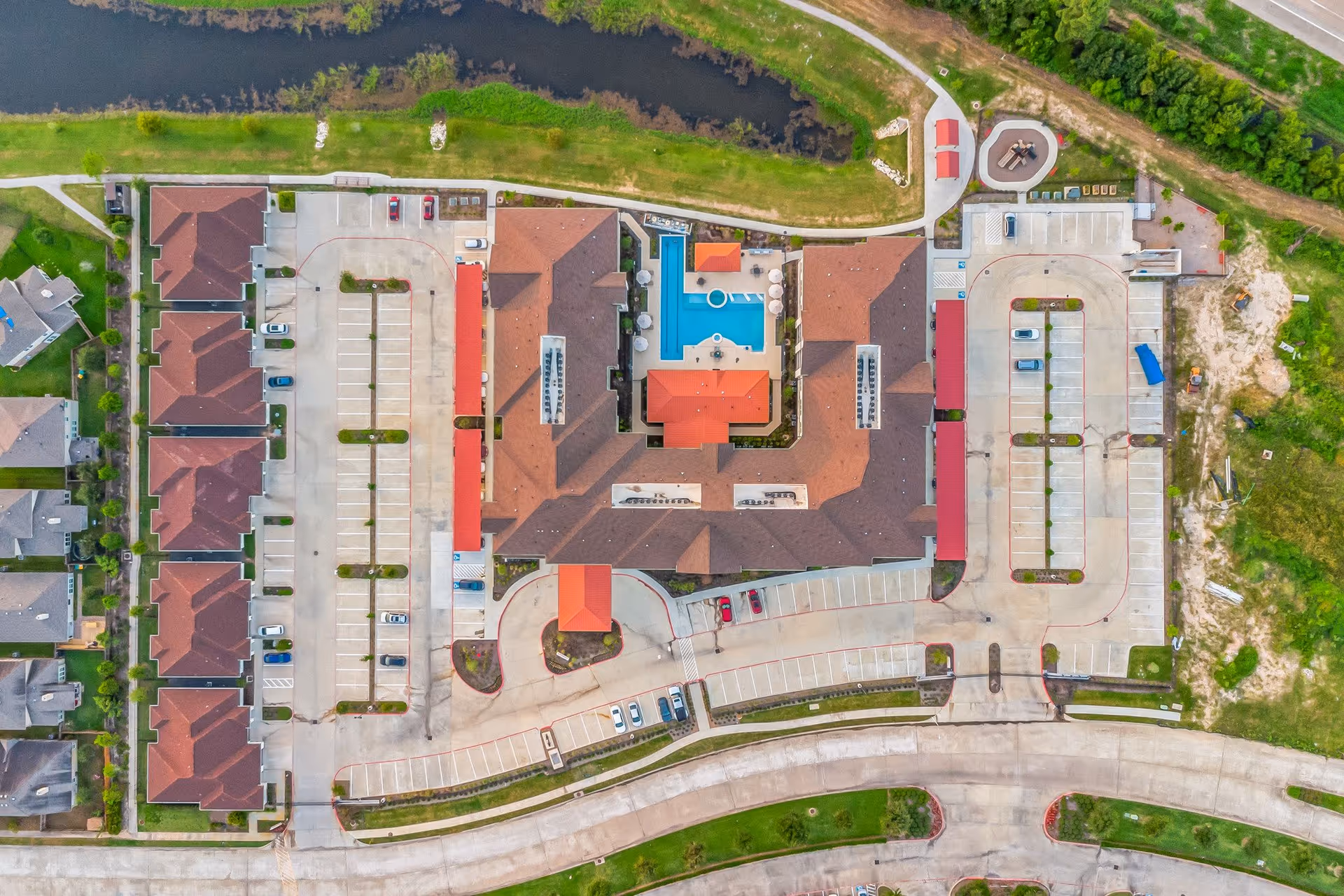 Aerial view of a senior living complex with a central building surrounding a courtyard pool, parking lots, driveways, and adjacent townhome-style units.