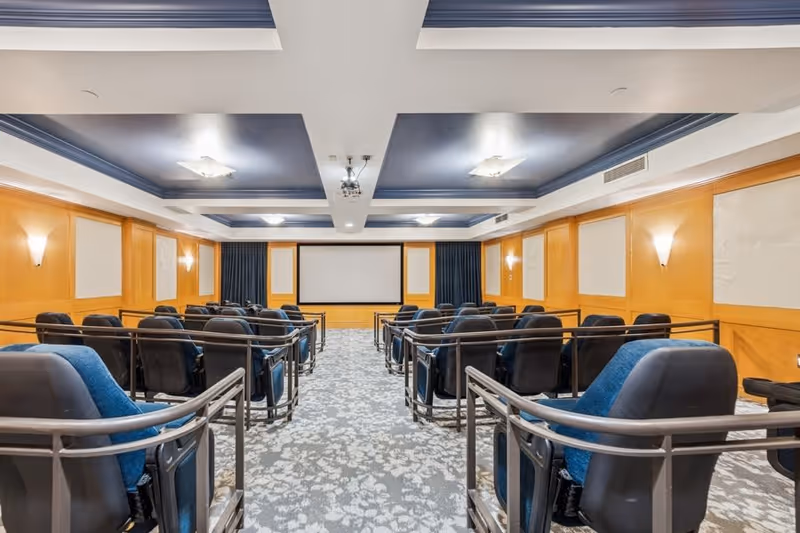 A small theater room with multiple rows of black and blue cushioned seats facing a large white projection screen. The room has wood-paneled walls, soft wall sconces for lighting, and a patterned carpet. The ceiling features recessed lighting and a projector mounted in the center.