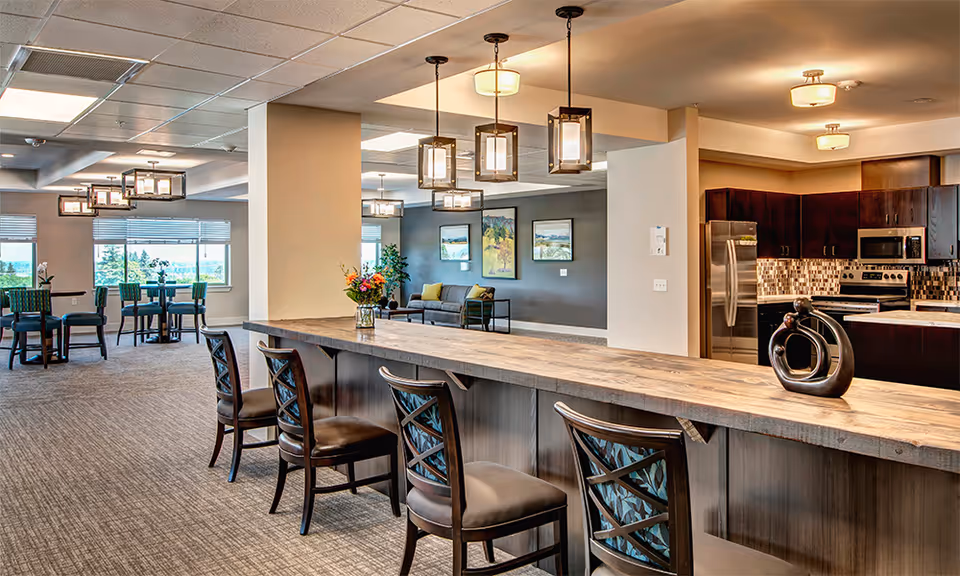 Spacious senior living community interior featuring a long wooden counter with four cushioned chairs, modern pendant lights hanging above, a kitchen area with stainless steel appliances and dark cabinetry, and a seating area with tables and chairs near large windows letting in natural light.