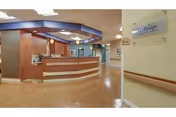Reception area inside a senior living facility with a wooden front desk, overhead lighting, and a hallway leading to other rooms. A sign on the wall reads 'Legacy Barberton'.
