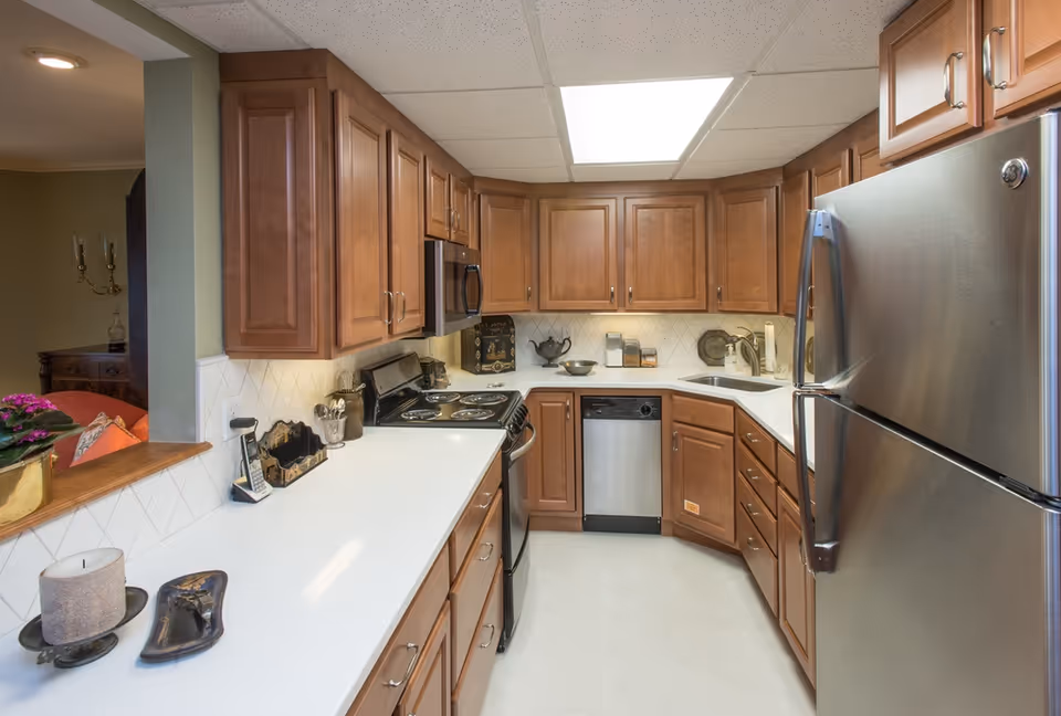 A modern kitchen with wooden cabinets, white countertops, and stainless steel appliances including a refrigerator, dishwasher, and stove. The kitchen has a tiled backsplash and a ceiling light panel. There is a small pass-through window opening to an adjacent room with a glimpse of a sofa and decorative items.