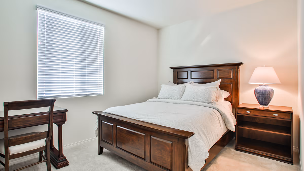 Neat bedroom with a wooden bed, matching nightstand and lamp, a small desk and chair beside a window with blinds.