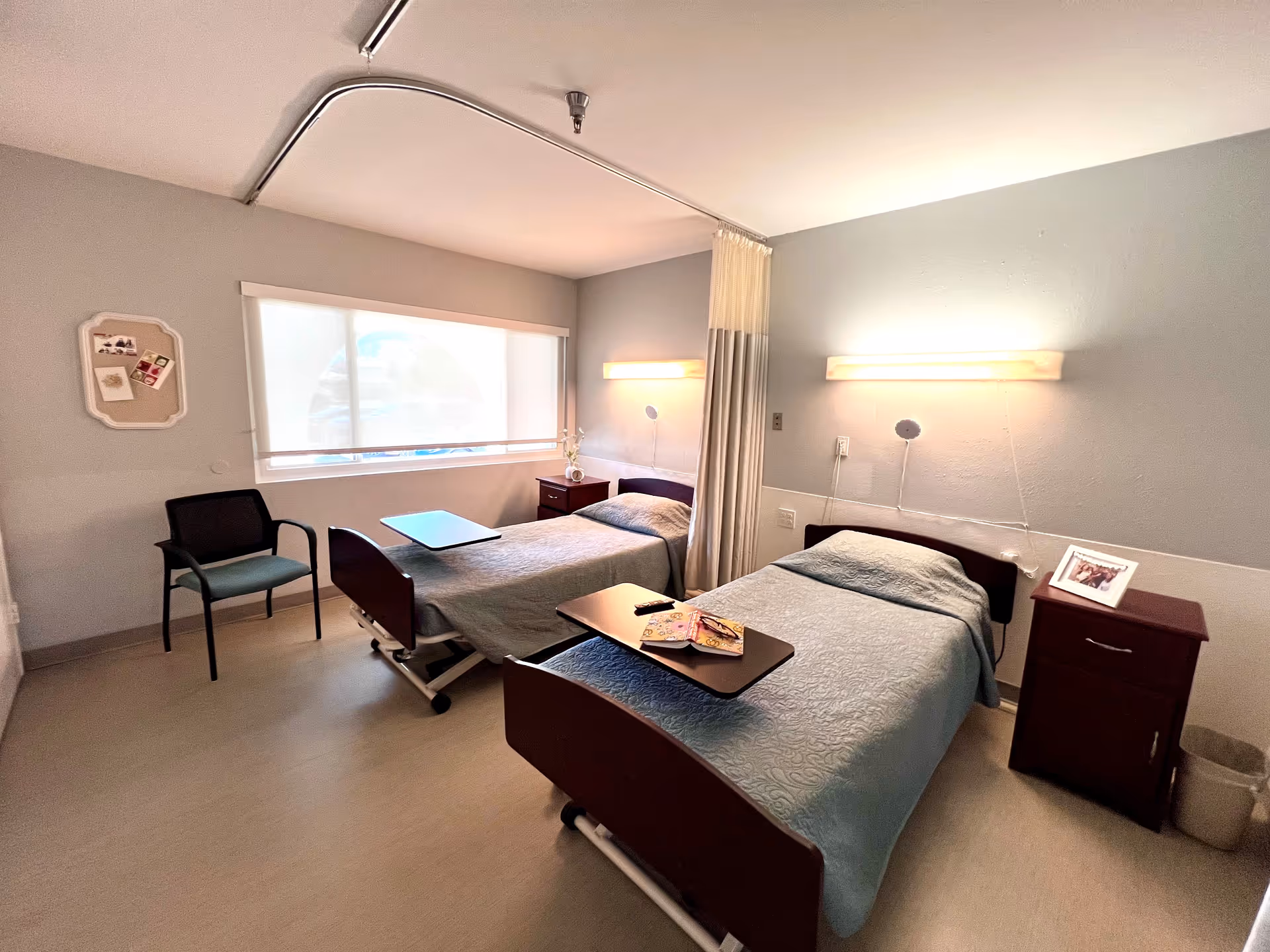 A bright and clean room with two single hospital-style beds separated by a privacy curtain. Each bed has a small overbed table, one with a book and remote control on it. There is a window with a roller shade, a chair, two bedside tables with a framed photo and a vase with flowers, and wall-mounted lights above each bed.