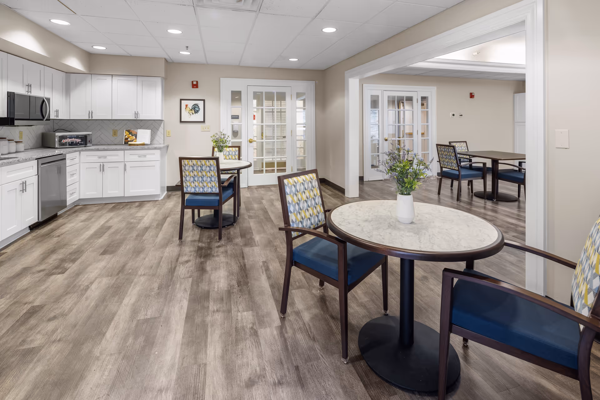 Bright communal dining area with round tables and chairs adjacent to a kitchenette with white cabinets and stainless appliances.
