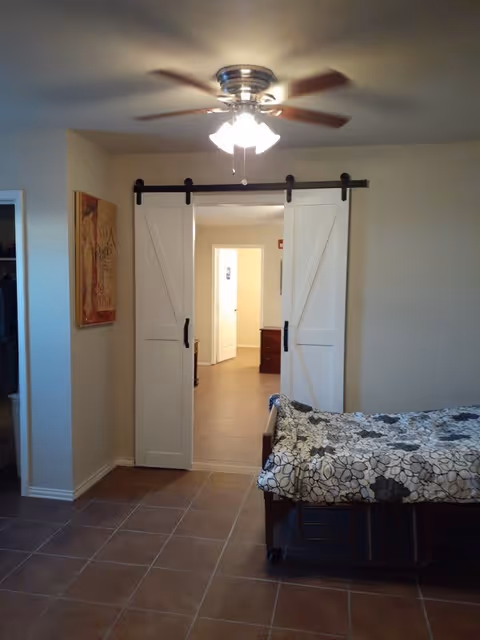 Interior view of a room with a ceiling fan light fixture, sliding barn doors on a black rail, a bed with floral patterned bedding, and a hallway leading to another room with an open door.