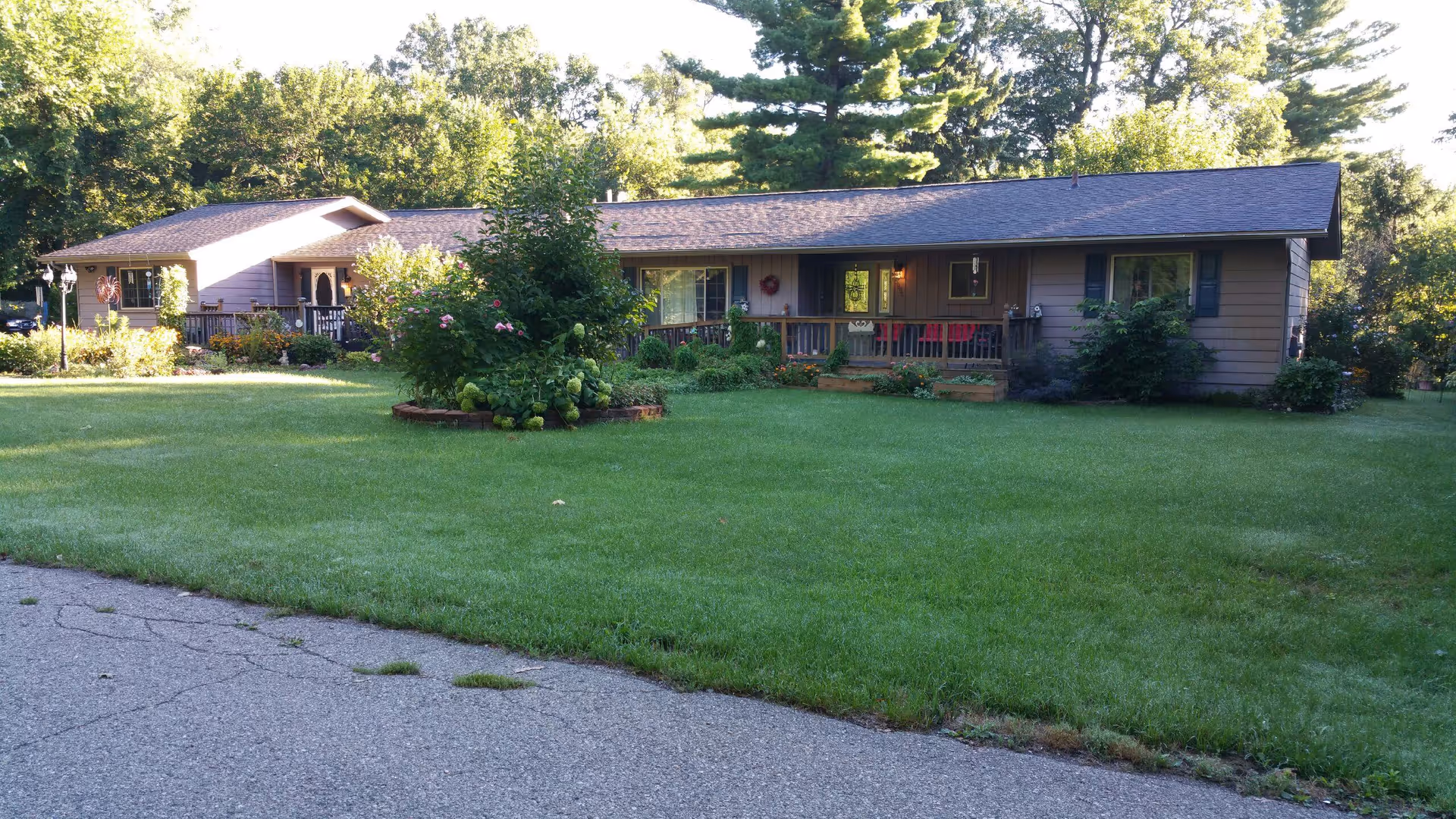 Single-story residential building with a large green lawn in front, surrounded by trees and bushes. The building has a porch with seating and is painted in a muted brown color with a dark roof.