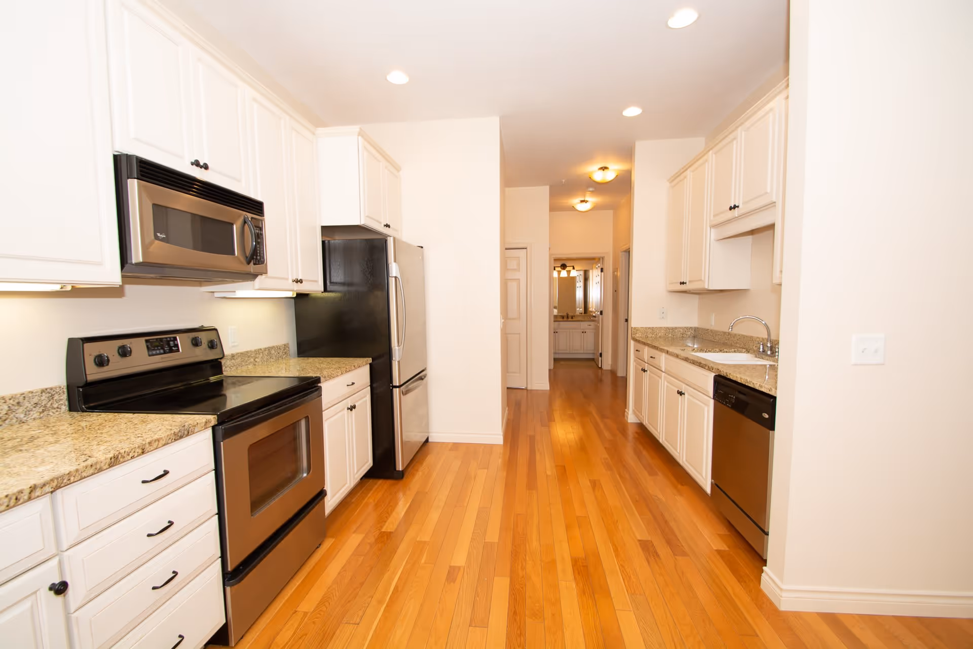 A bright kitchen with wooden floors, white cabinets, granite countertops, and stainless steel appliances including a microwave, stove, refrigerator, and dishwasher. The kitchen has recessed lighting and a hallway leading to another room in the background.