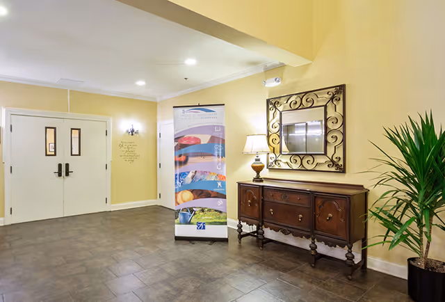 A well-lit interior lobby with double doors, a decorative console table topped with a lamp and mirror, a potted plant, and a promotional banner.