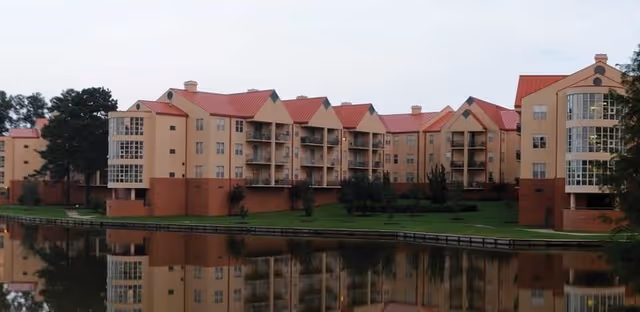 Exterior view of a multi-story residential building with red roofs and beige walls, reflected in a calm body of water in front of it. The building is surrounded by green grass and trees.