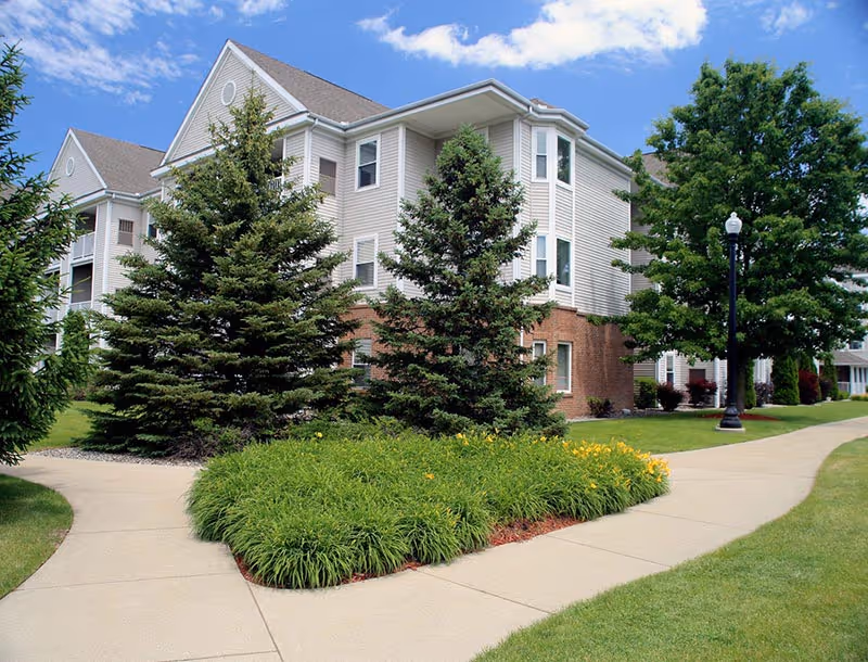 Exterior view of a multi-story residential building with beige siding and brick accents, surrounded by green trees, shrubs, and well-maintained landscaping. A concrete walkway curves through the grass and flower beds under a partly cloudy blue sky.