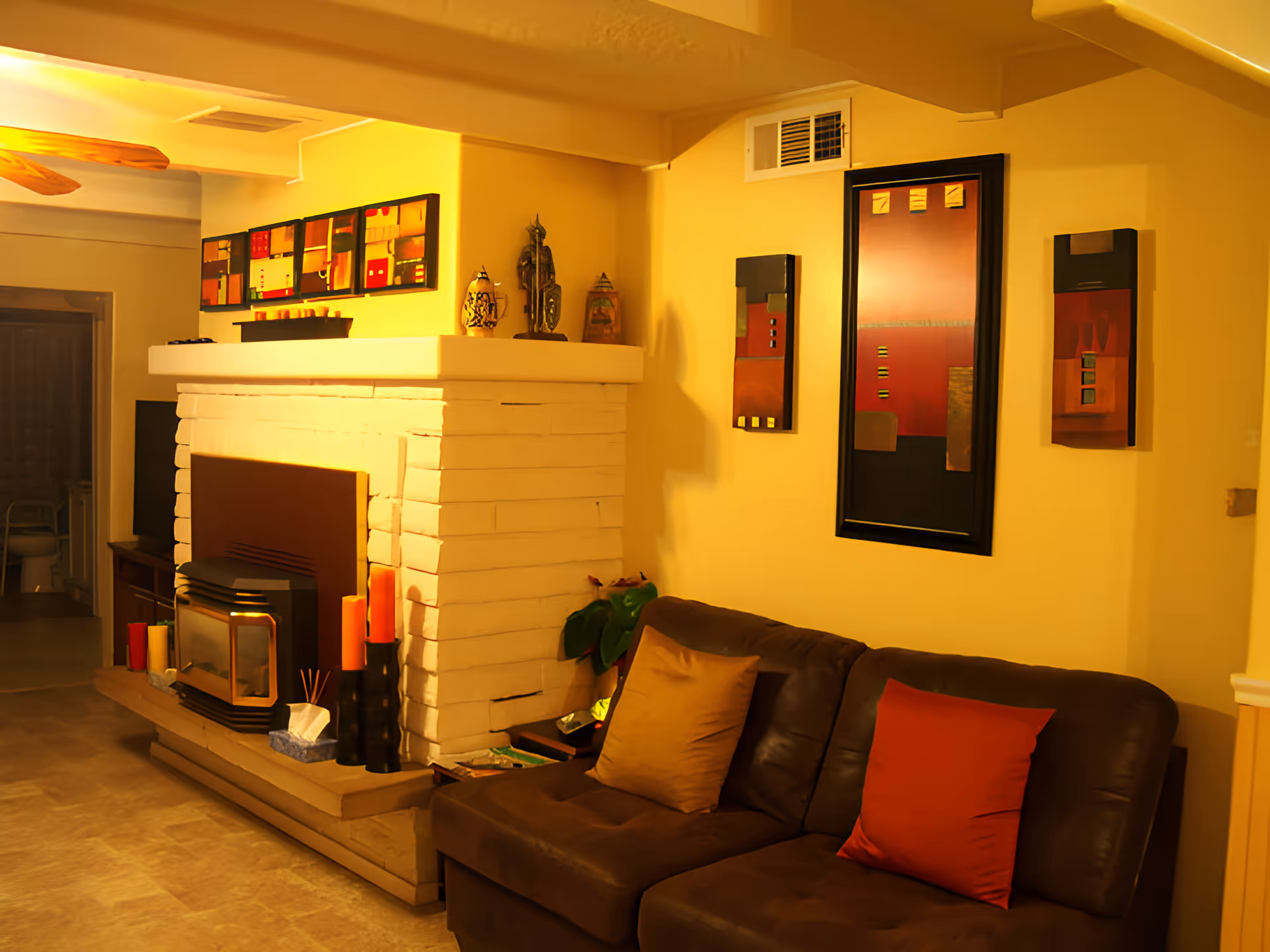 Cozy living room with a brown leather couch adorned with red and beige pillows, a white brick fireplace with decorative items and candles on the mantel, and abstract artwork hanging on the yellow walls.