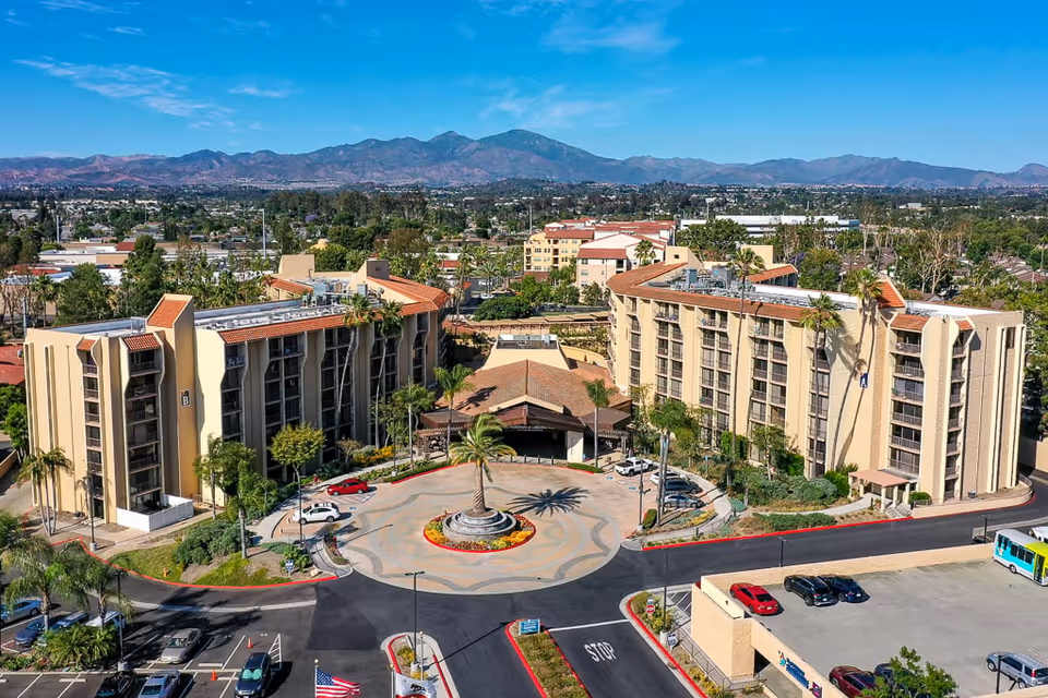 Aerial view of Meridian at Laguna Hills senior living facility showing two large beige multi-story buildings with red-tiled roofs surrounding a circular driveway with a central palm tree and landscaped flower bed. The facility is set against a backdrop of a sprawling suburban area with mountains in the distance under a clear blue sky.