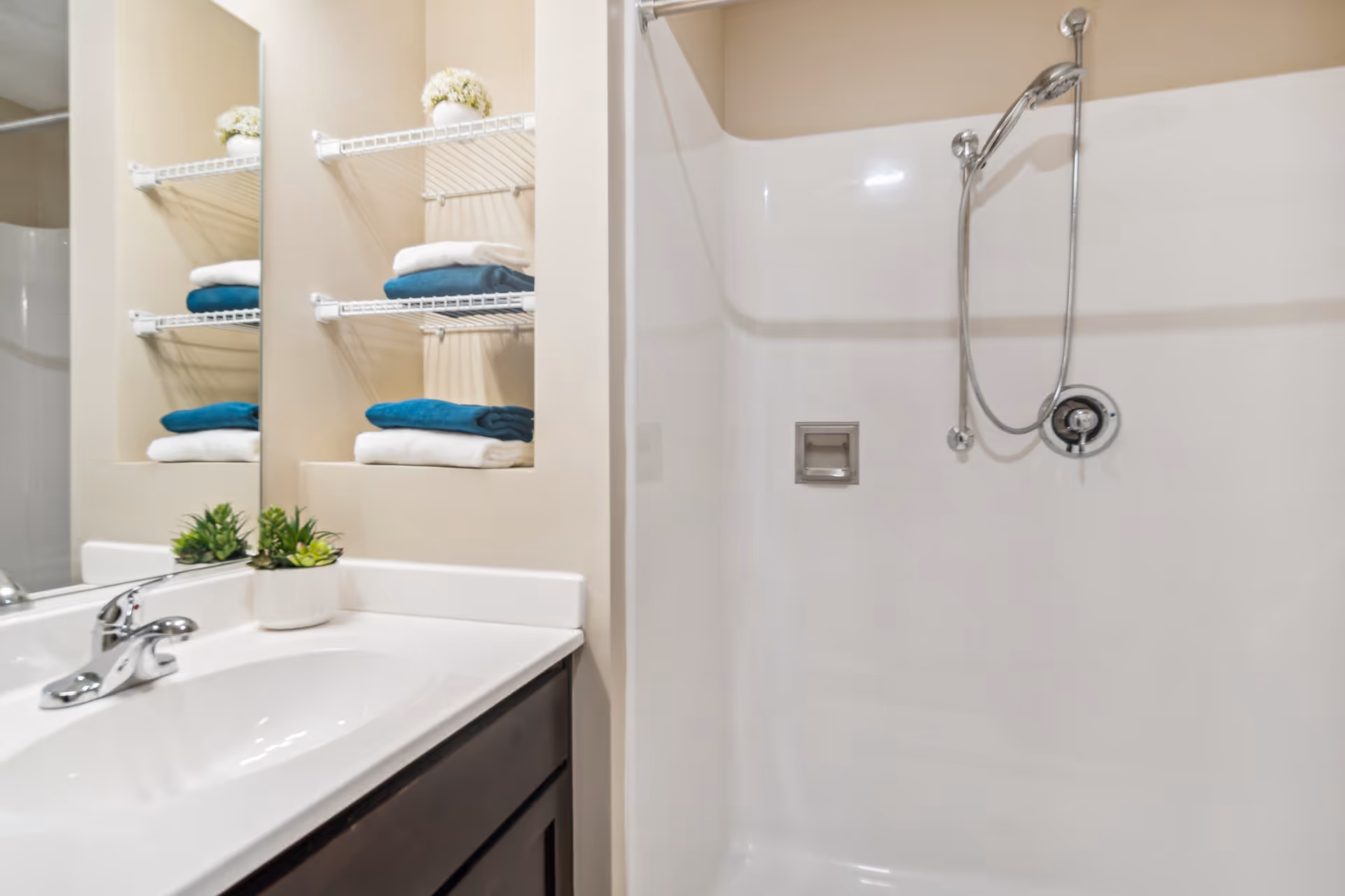A clean bathroom featuring a white sink with a silver faucet, a mirror above the sink, and a shower area with a handheld showerhead. There are white and teal towels neatly folded on white wire shelves, along with small potted plants for decoration.
