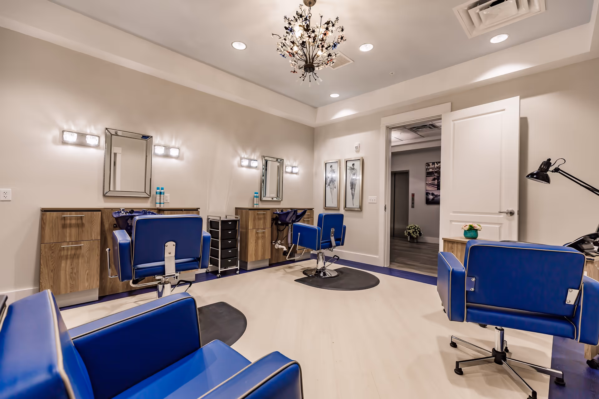Interior view of a salon area with blue salon chairs, mirrors, wooden cabinets, and bright lighting fixtures on the walls and ceiling. The room has light-colored flooring and a decorative chandelier, with an open door leading to another room.